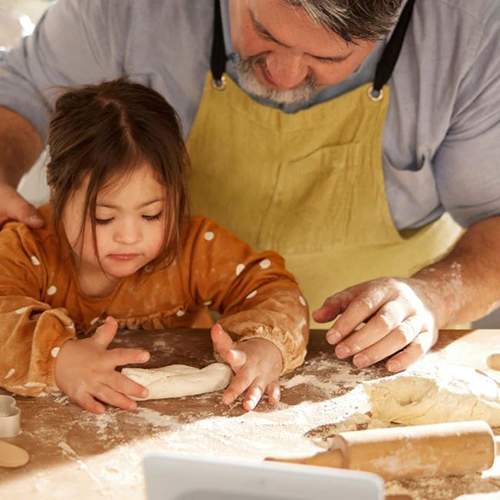 An overhead shot of the father's hands guiding his daughter as she learns to flatten dough with a rolling pin.